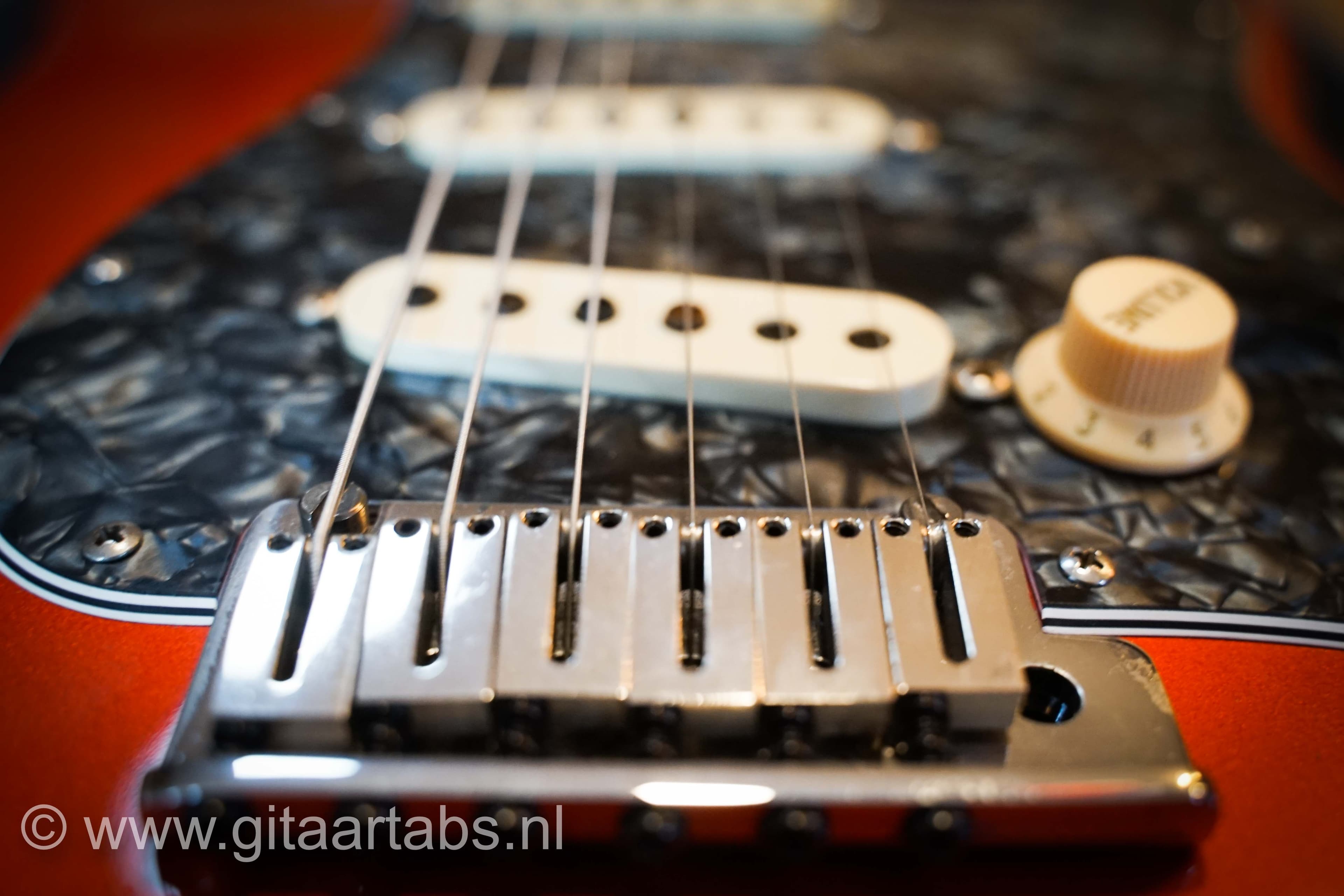 Close-up van een elektrische gitaar met metalen snaren en een zwartgemêleerd slagplaat. De gitaar is rood. Tekst: gitaartabs.nl.