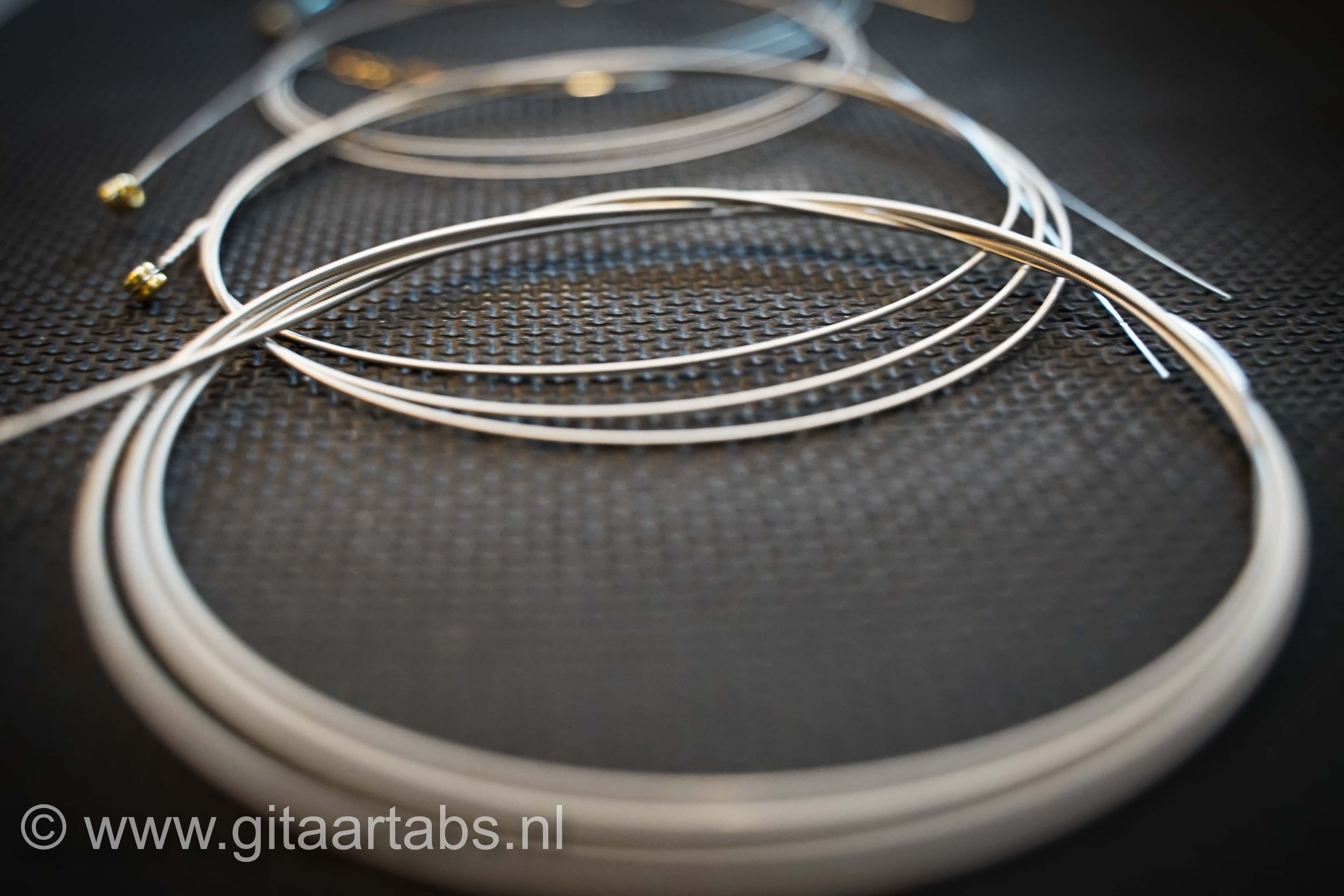 Gitaar snaren liggen op een zwarte getextureerde ondergrond. Tekst onderaan: www.gitaartabs.nl. Rustige, technische sfeer.